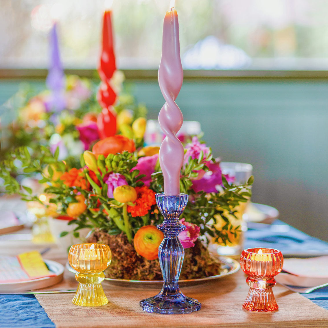 Colorful table setting with floral arrangement, candles, and glass holders on a wooden table.